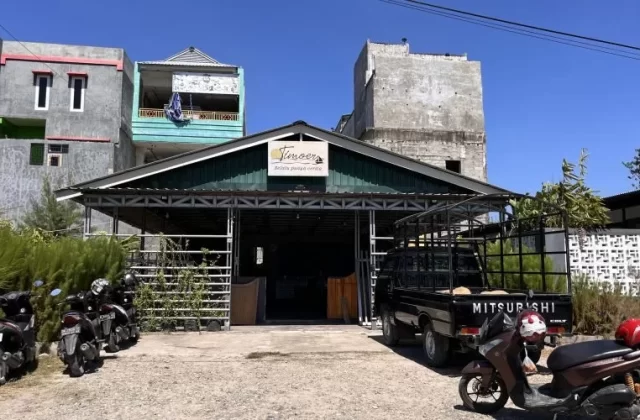 Timoer Cafe, a building with a dark green roof, stands under a clear blue sky with a black pickup truck and motorbikes parked outside.