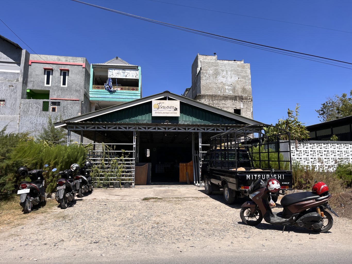 Timoer Cafe, a building with a dark green roof, stands under a clear blue sky with a black pickup truck and motorbikes parked outside.