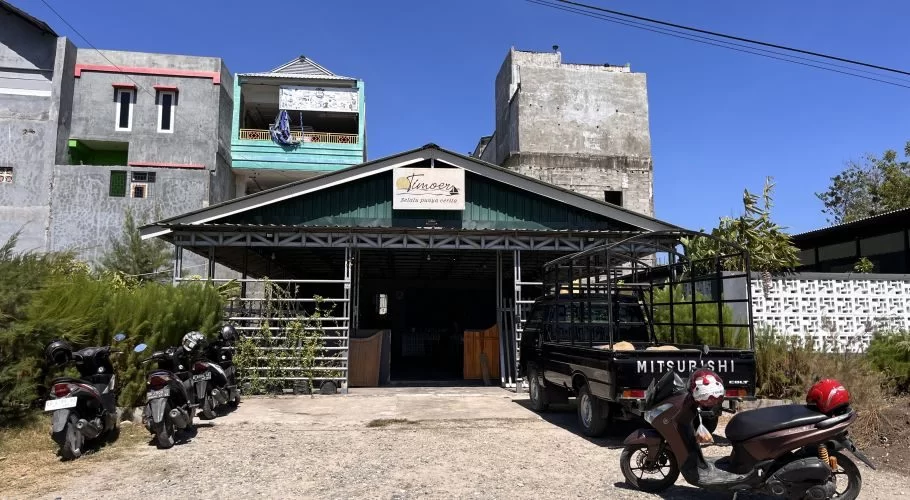 Timoer Cafe, a building with a dark green roof, stands under a clear blue sky with a black pickup truck and motorbikes parked outside.