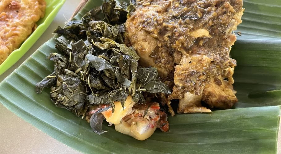 Spiced chicken, likely Ayam Betutu, is served with dark green leafy vegetables on a vibrant banana leaf, with a blurry fried item in the background.