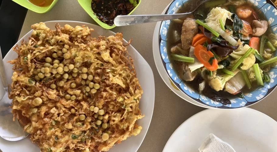 A large golden-brown omelet topped with green peas sits next to a bowl of colorful Capcay stir-fry with vegetables and shrimp on a table.