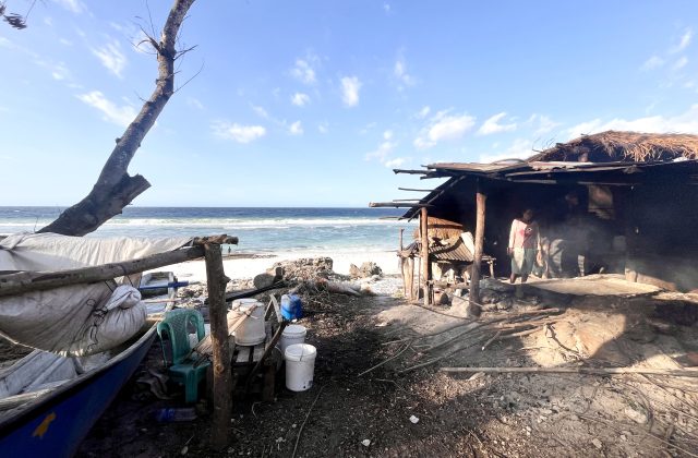 A person stands inside a rustic, open-sided hut on a sandy beach, with smoke rising from a traditional salt-making fire near the ocean under a blue sky.