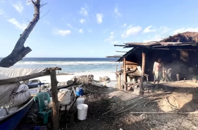 A person stands inside a rustic, open-sided hut on a sandy beach, with smoke rising from a traditional salt-making fire near the ocean under a blue sky.