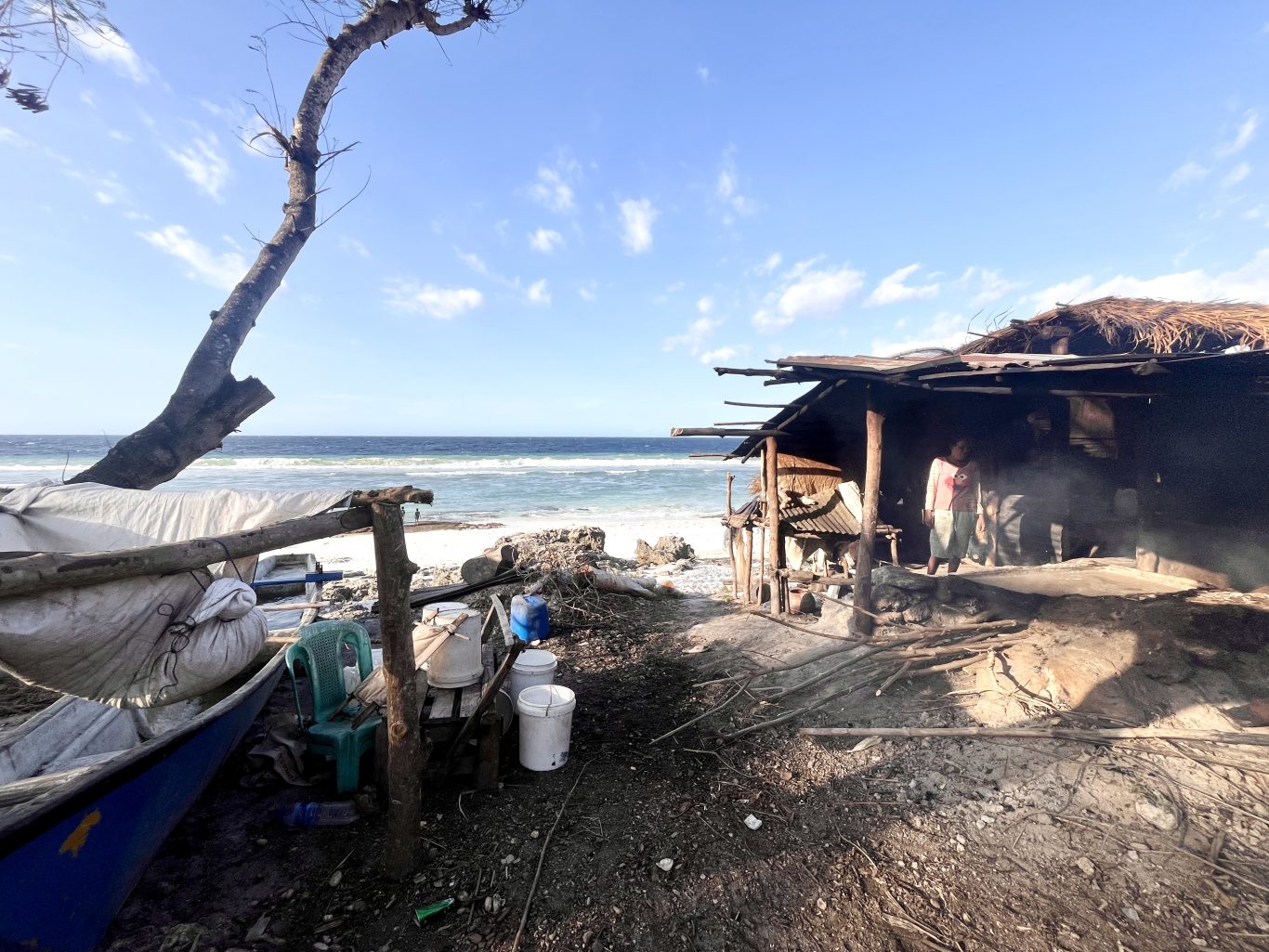 A person stands inside a rustic, open-sided hut on a sandy beach, with smoke rising from a traditional salt-making fire near the ocean under a blue sky.