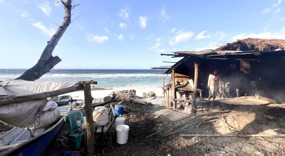 A person stands inside a rustic, open-sided hut on a sandy beach, with smoke rising from a traditional salt-making fire near the ocean under a blue sky.