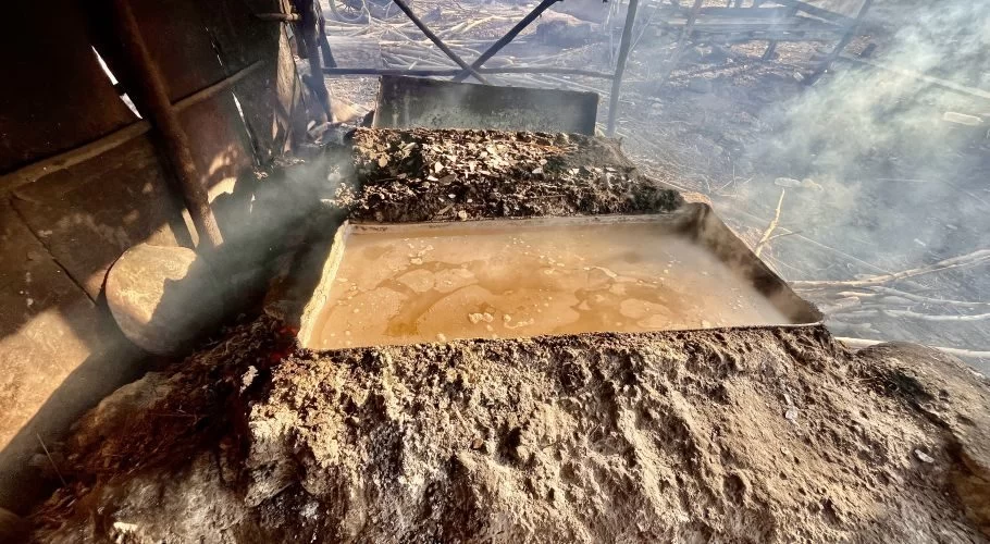 Seawater boils in a rectangular metal pan, surrounded by dark soil and emitting steam, as part of a traditional salt-making process.