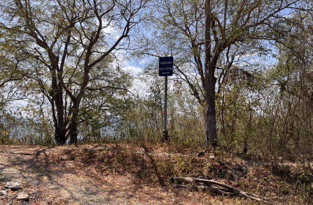 A blue sign for "KAMPUNG UBU BEWI" with a left arrow stands on a dirt path surrounded by dry trees and brush under a bright blue sky.