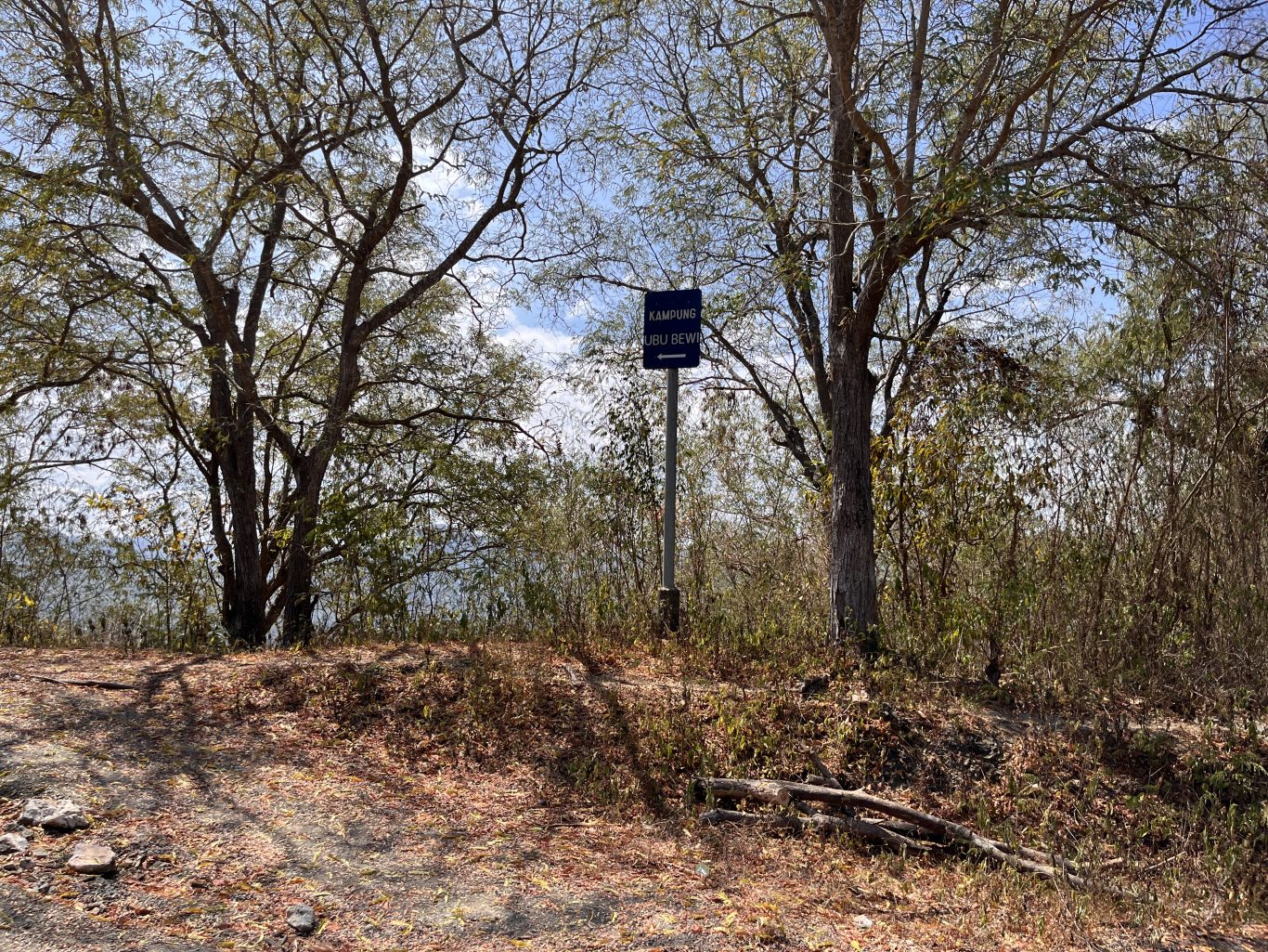 A blue sign for "KAMPUNG UBU BEWI" with a left arrow stands on a dirt path surrounded by dry trees and brush under a bright blue sky.