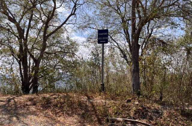 A blue sign for "KAMPUNG UBU BEWI" with a left arrow stands on a dirt path surrounded by dry trees and brush under a bright blue sky.