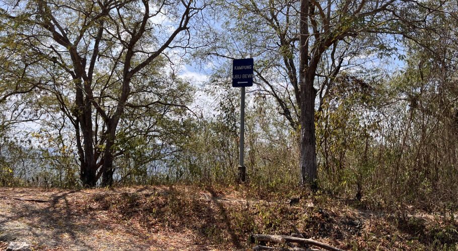 A blue sign for "KAMPUNG UBU BEWI" with a left arrow stands on a dirt path surrounded by dry trees and brush under a bright blue sky.