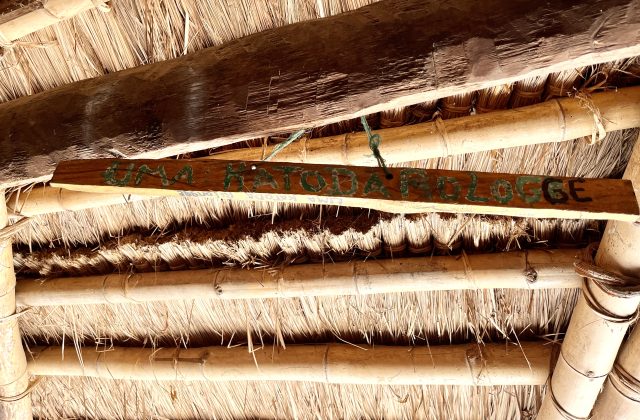 A wooden sign with "UMA KATODA ROLOGGE" painted in green and black hangs from a traditional thatched roof supported by bamboo and wood beams.