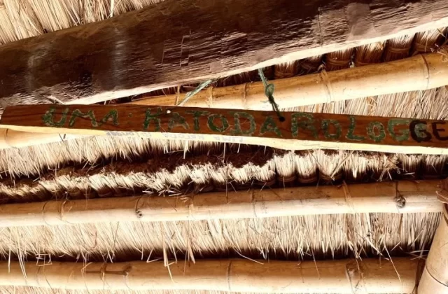 A wooden sign with "UMA KATODA ROLOGGE" painted in green and black hangs from a traditional thatched roof supported by bamboo and wood beams.