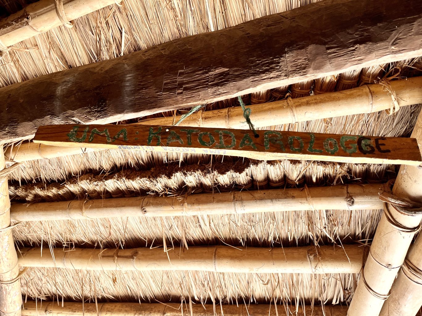 A wooden sign with "UMA KATODA ROLOGGE" painted in green and black hangs from a traditional thatched roof supported by bamboo and wood beams.