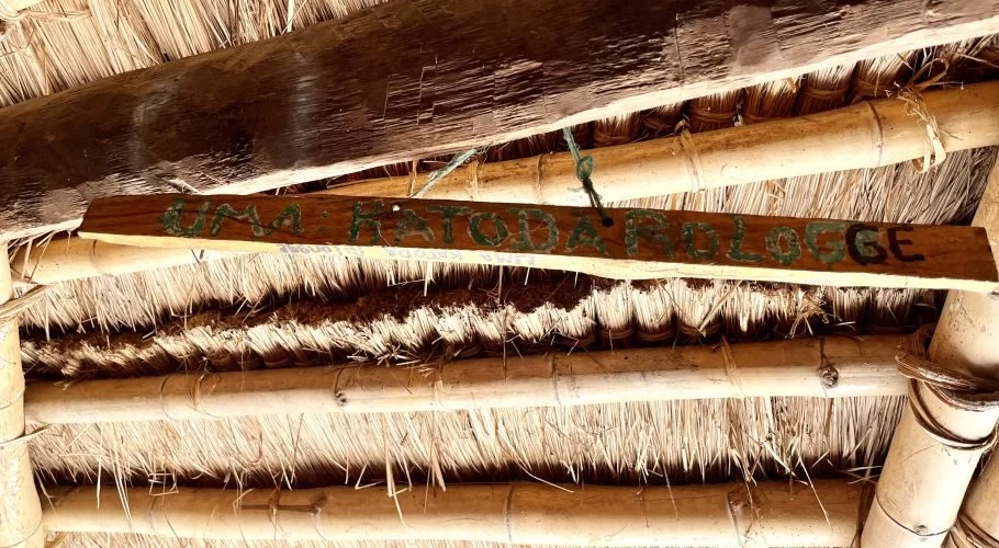 A wooden sign with "UMA KATODA ROLOGGE" painted in green and black hangs from a traditional thatched roof supported by bamboo and wood beams.