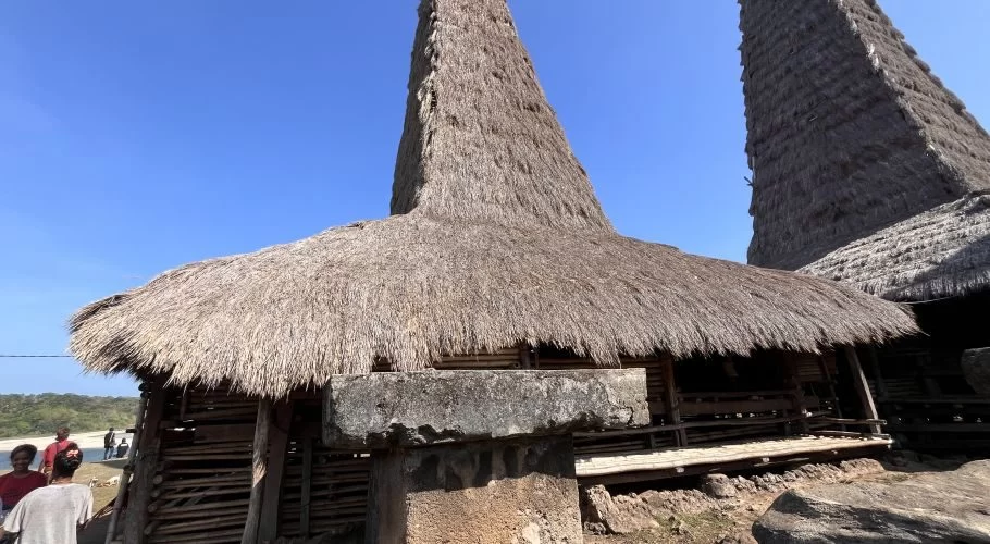 Two traditional Sumba houses with tall, conical thatched roofs stand under a clear blue sky, with people and a body of water in the background.