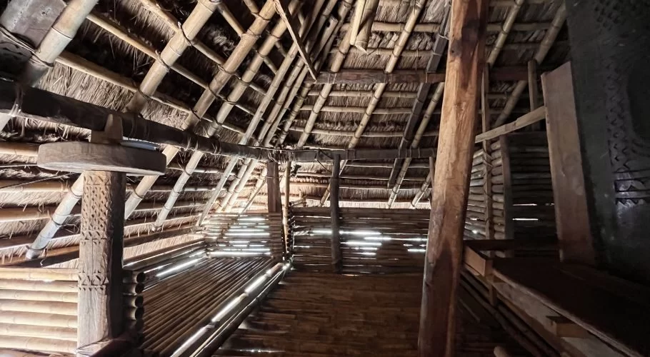 Interior view of a traditional Sumba house with a high thatched roof, sturdy wooden beams, and a carved central pillar, illuminated by sunlight.