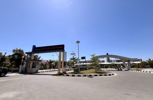 Umbu Mehang Kunda Airport entrance in East Sumba has a large sign, security booth, and modern arched structure under a clear blue sky.