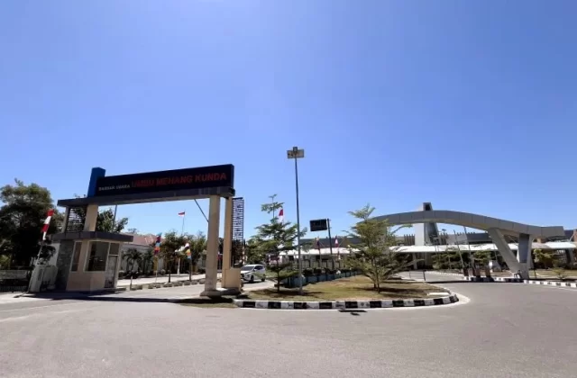 Umbu Mehang Kunda Airport entrance in East Sumba has a large sign, security booth, and modern arched structure under a clear blue sky.