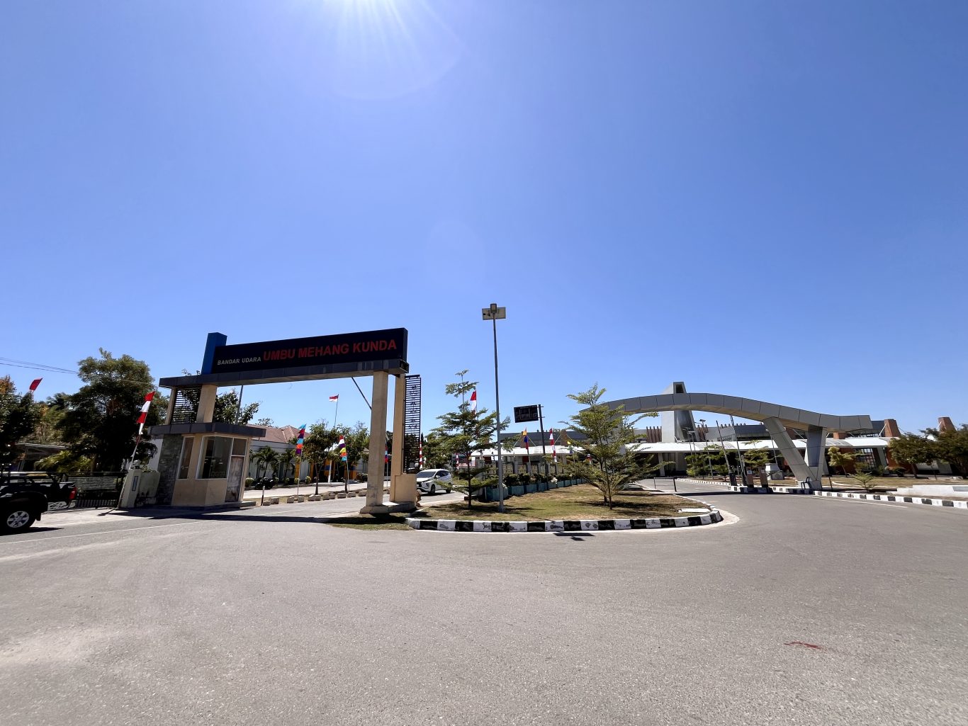 Umbu Mehang Kunda Airport entrance in East Sumba has a large sign, security booth, and modern arched structure under a clear blue sky.