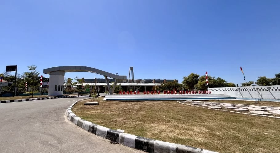 The entrance to Umbu Mehang Kunda Airport features a curving road, a large arched gate, and a red sign against a clear blue sky.