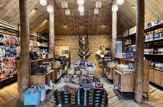 The interior of a Sumba souvenir shop features bamboo walls and ceiling, wooden pillars, and displays of colorful ikat fabrics, woven bags, and traditional artifacts.