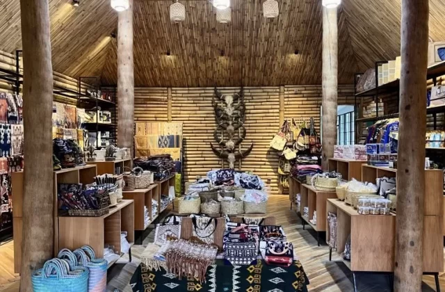 The interior of a Sumba souvenir shop features bamboo walls and ceiling, wooden pillars, and displays of colorful ikat fabrics, woven bags, and traditional artifacts.