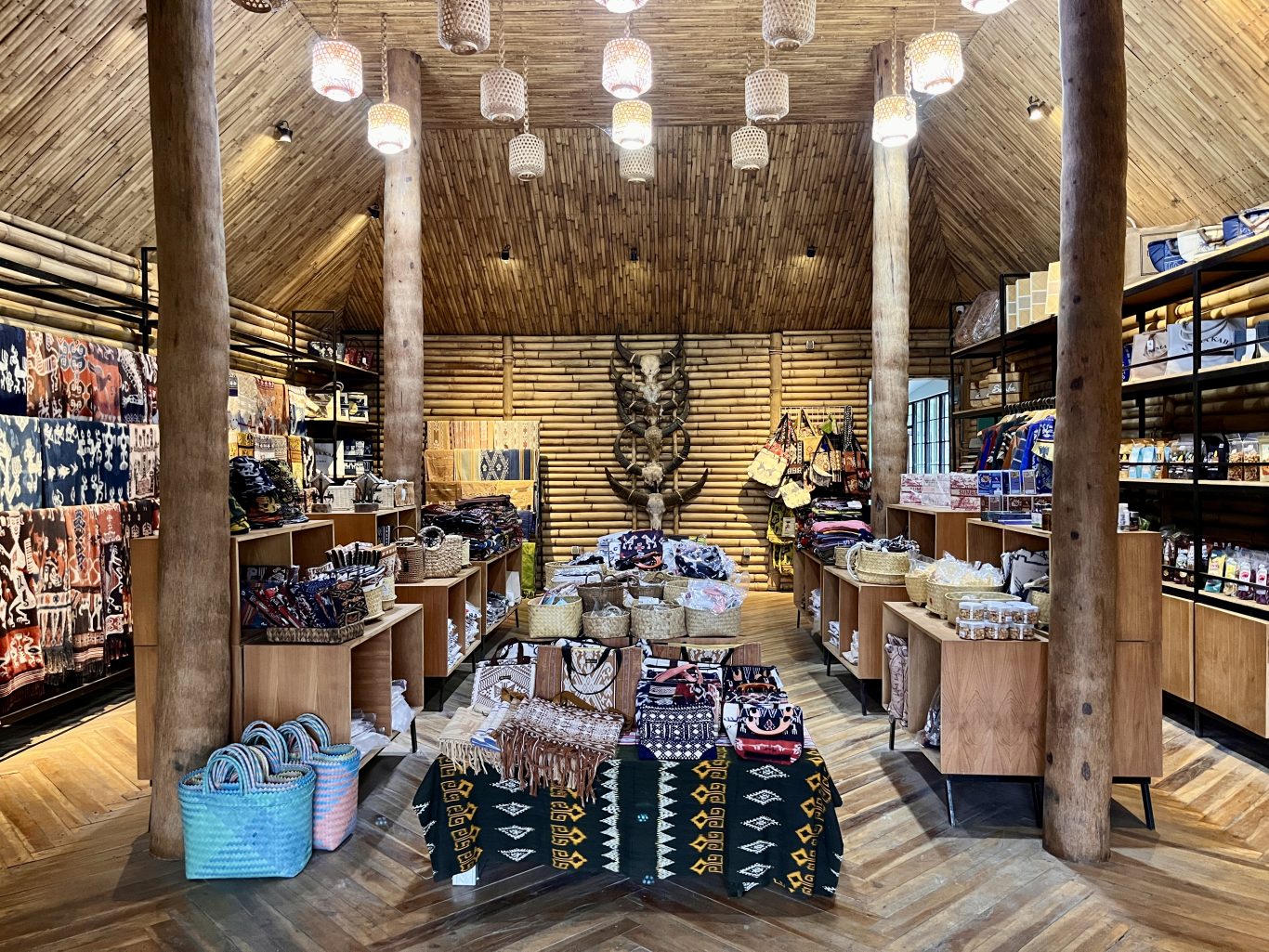 The interior of a Sumba souvenir shop features bamboo walls and ceiling, wooden pillars, and displays of colorful ikat fabrics, woven bags, and traditional artifacts.