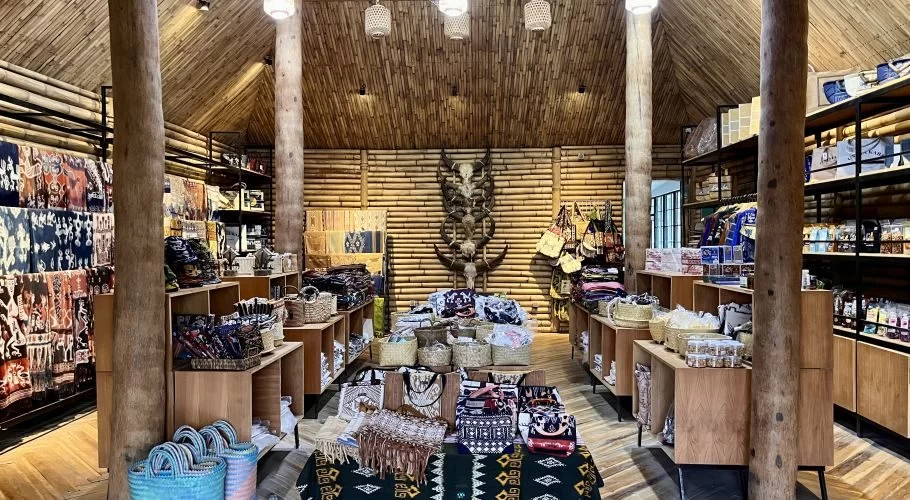 The interior of a Sumba souvenir shop features bamboo walls and ceiling, wooden pillars, and displays of colorful ikat fabrics, woven bags, and traditional artifacts.