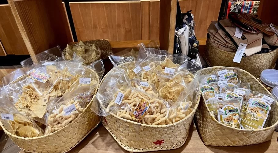 Various local Sumba snacks, including chips, twisted crackers, and egg peanuts, are displayed in three light brown woven baskets on a wooden shelf.
