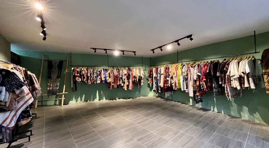 A spacious souvenir shop interior features light green walls, grey tiled floors, and multiple racks displaying colorful clothing with Sumba motifs, illuminated by ceiling spotlights.