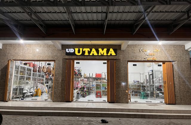 The brightly lit exterior of UD Utama, a souvenir shop in Waingapu, features a brown tiled facade and three glass display sections at night.