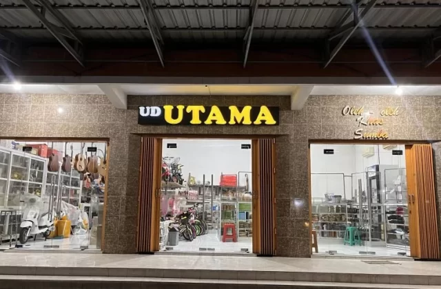 The brightly lit exterior of UD Utama, a souvenir shop in Waingapu, features a brown tiled facade and three glass display sections at night.
