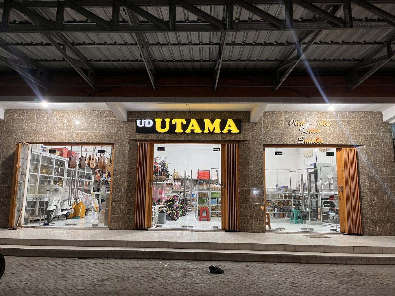 The brightly lit exterior of UD Utama, a souvenir shop in Waingapu, features a brown tiled facade and three glass display sections at night.