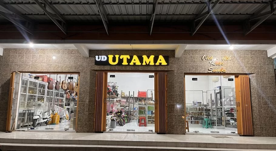 The brightly lit exterior of UD Utama, a souvenir shop in Waingapu, features a brown tiled facade and three glass display sections at night.