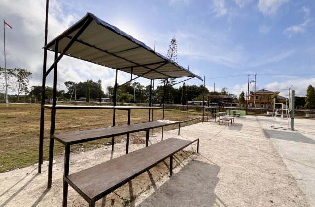 Empty wooden spectator benches under a white canopy overlook a concrete volleyball court and dry grassy field in West Sumba, Indonesia, under a bright sky.