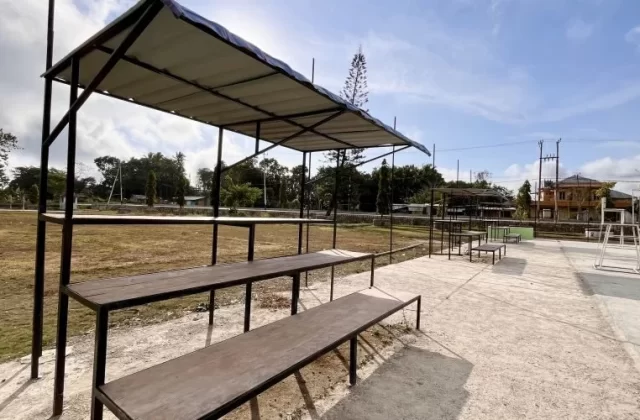 Empty wooden spectator benches under a white canopy overlook a concrete volleyball court and dry grassy field in West Sumba, Indonesia, under a bright sky.