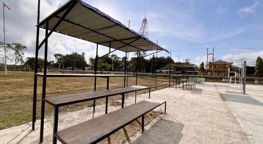 Empty wooden spectator benches under a white canopy overlook a concrete volleyball court and dry grassy field in West Sumba, Indonesia, under a bright sky.