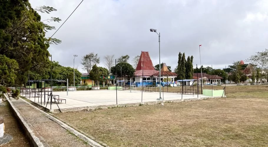 An empty outdoor volleyball court with concrete surface and dry grass, surrounded by green trees and buildings with traditional red roofs under a cloudy sky in Loli.