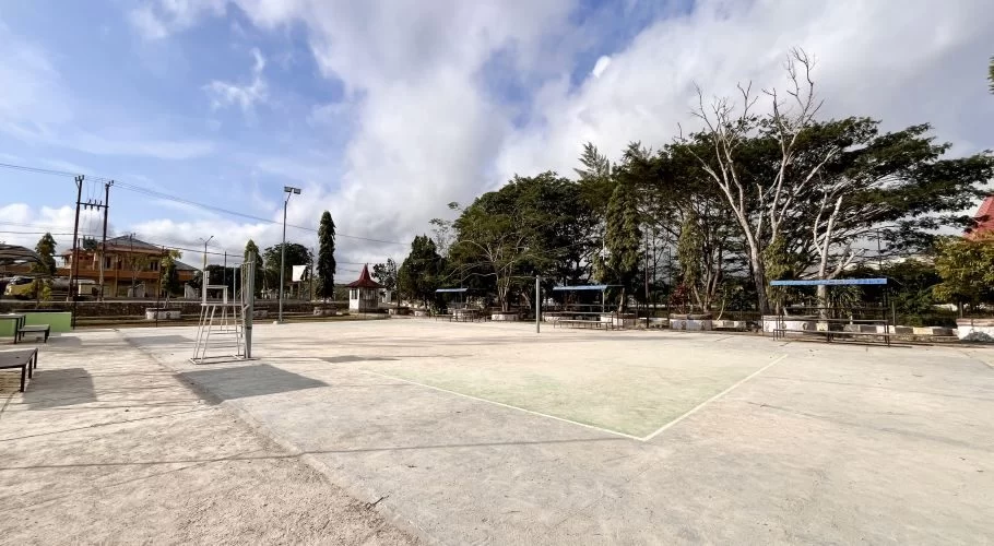 A wide view of an empty outdoor concrete volleyball court with a green playing area, surrounded by trees, spectator seating, and distant buildings under a partly cloudy sky in Loli, West Sumba.