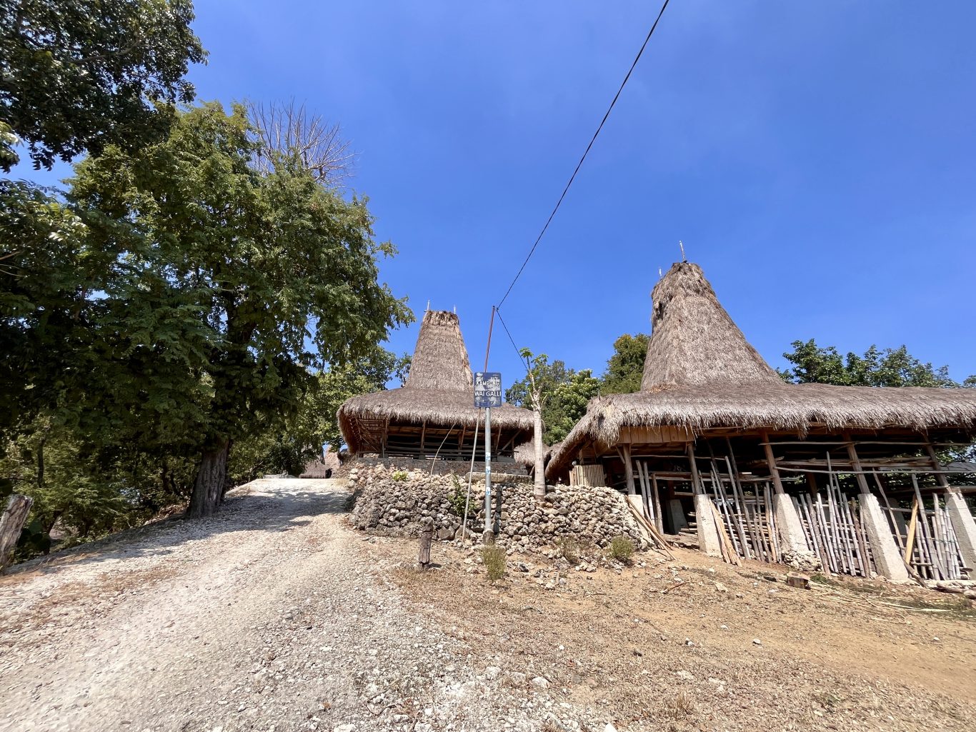 Two traditional Sumbanese houses with tall, conical thatched roofs stand beside a dirt path under a clear blue sky, surrounded by green trees.