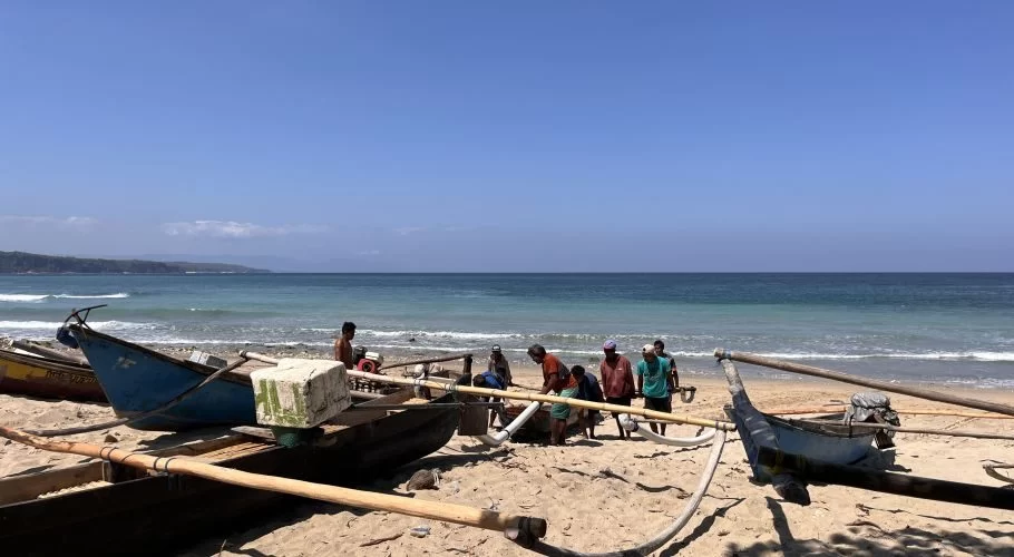 Several fishermen work around blue outrigger boats on a sunny sandy beach with a clear blue ocean and distant green coastline under a bright sky.