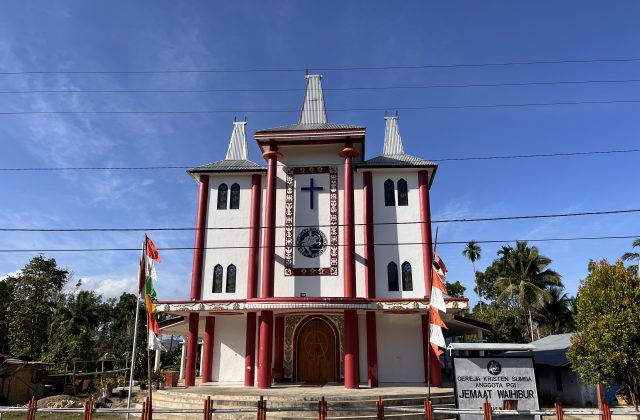 Waihibur Church, a white building with maroon pillars and three tall, Sumba-style roofs, stands under a blue sky, flanked by green trees and flags.