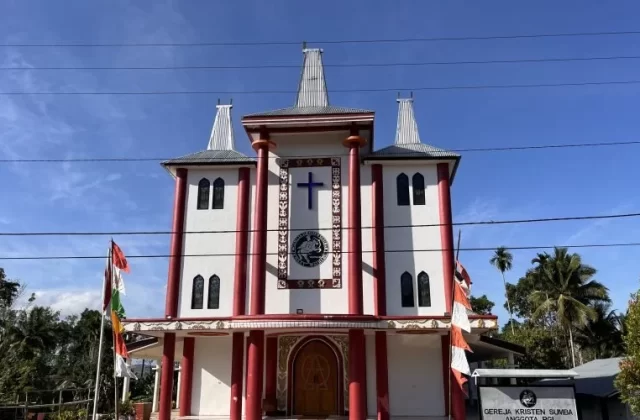 Waihibur Church, a white building with maroon pillars and three tall, Sumba-style roofs, stands under a blue sky, flanked by green trees and flags.