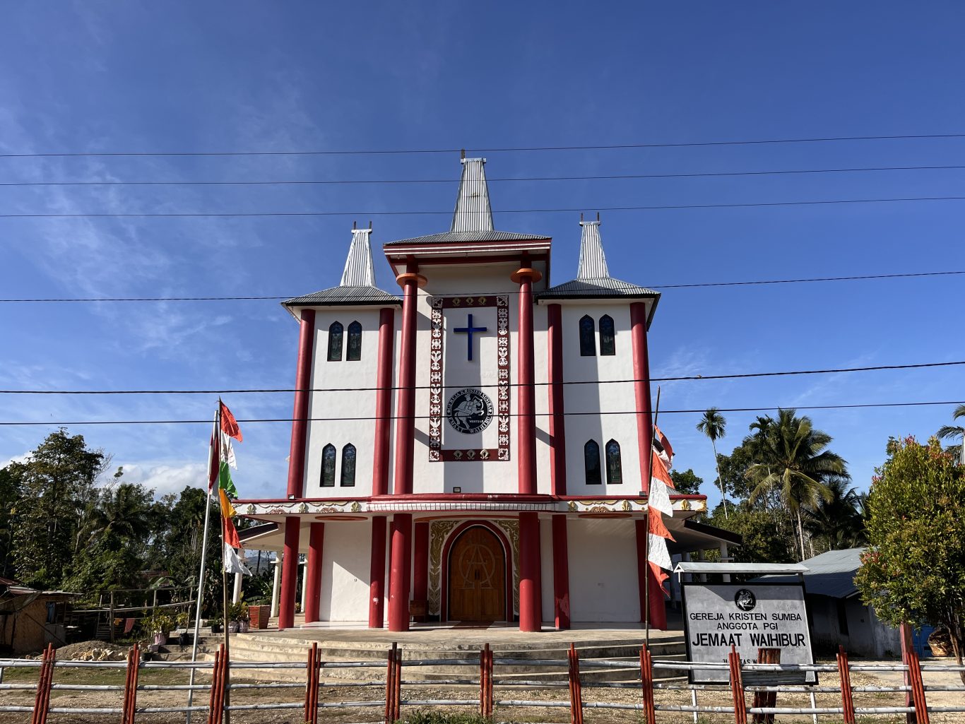 Waihibur Church, a white building with maroon pillars and three tall, Sumba-style roofs, stands under a blue sky, flanked by green trees and flags.
