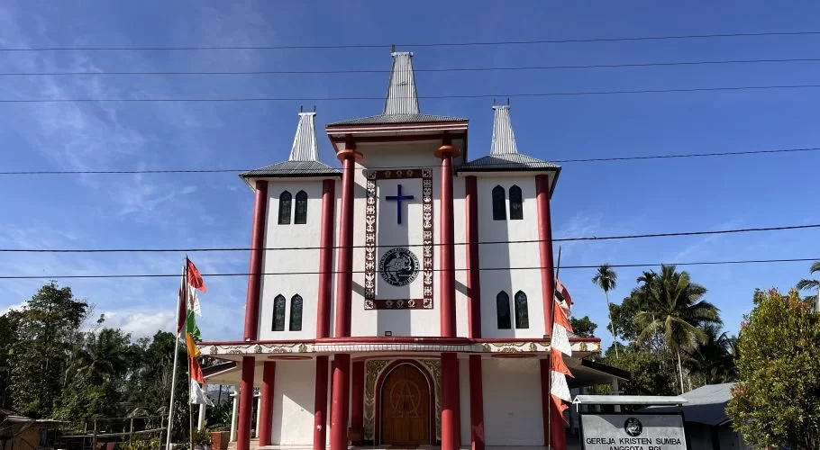 Waihibur Church, a white building with maroon pillars and three tall, Sumba-style roofs, stands under a blue sky, flanked by green trees and flags.