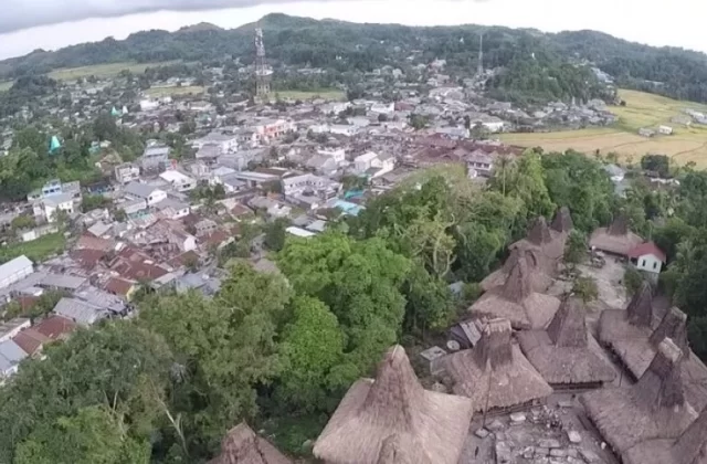 Traditional Sumba houses with tall thatched roofs stand in the foreground, backed by the modern town of Waikabubak and green hills under an overcast sky.