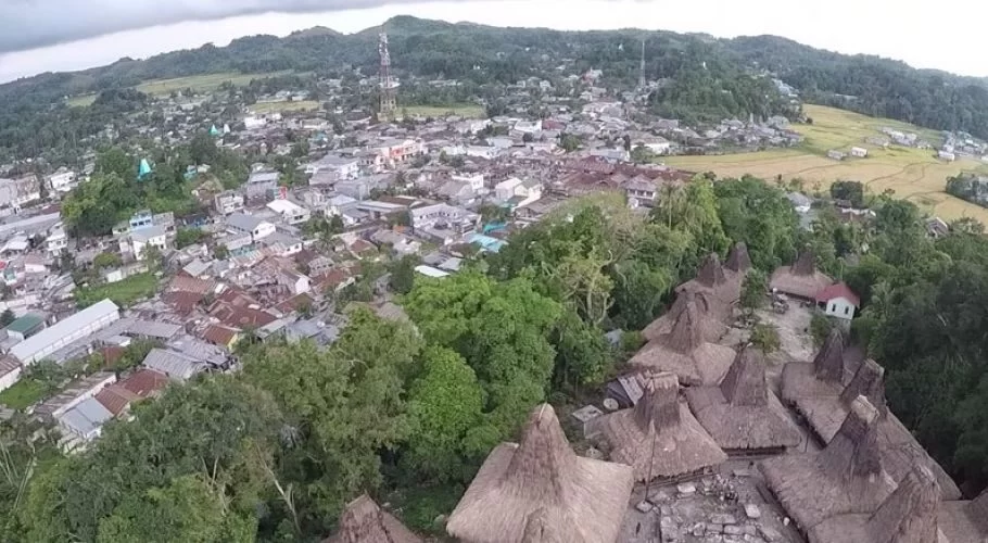 Traditional Sumba houses with tall thatched roofs stand in the foreground, backed by the modern town of Waikabubak and green hills under an overcast sky.