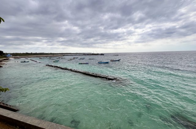 Waikelo Beach features clear blue-green water with numerous small boats floating near a long wooden pier under a cloudy sky.