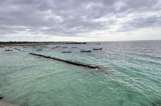 Waikelo Beach features clear blue-green water with numerous small boats floating near a long wooden pier under a cloudy sky.