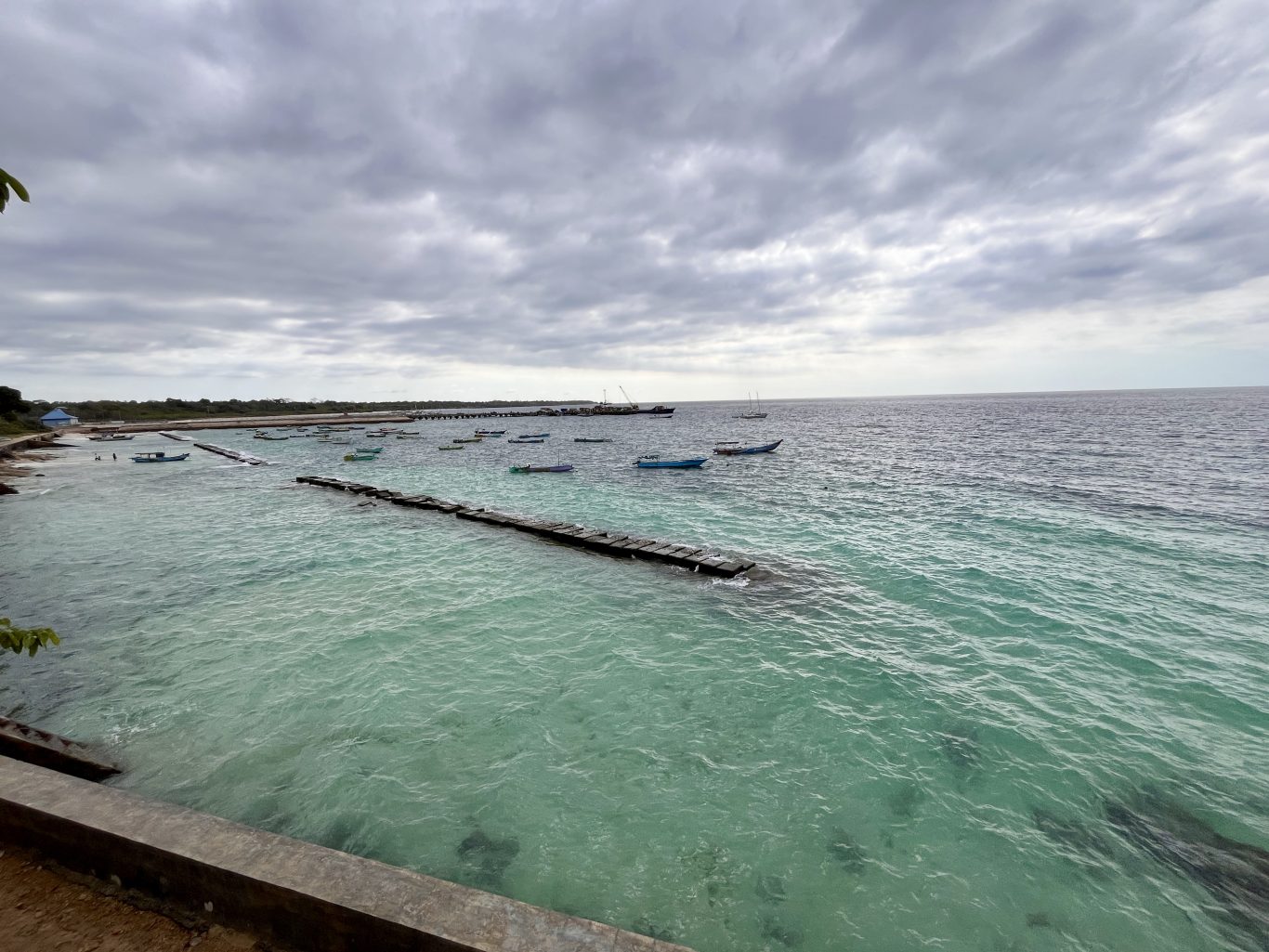 Waikelo Beach features clear blue-green water with numerous small boats floating near a long wooden pier under a cloudy sky.
